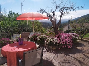 En patio med et bord og stole, en rød parasol, samt en have med blomster og et træ.