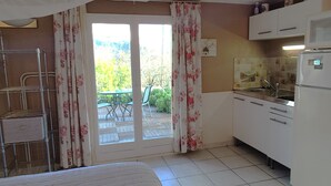 A compact kitchen with a refrigerator, sink, and cabinets. There is a sliding glass door leading to an outdoor patio with a table and chairs. - Studio (Bendejun)