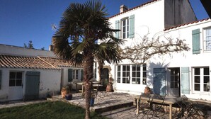 A white building with blue shutters, a stone patio, and a palm tree. - Au Coeur du Banzeau (Noirmoutier-en-l'Île)