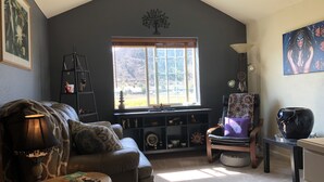 Un salon chaleureux avec un plafond haut, une grande fenêtre donnant sur un paysage, un canapé confortable, un fauteuil de lecture et une petite table avec une lampe et des livres.