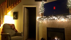 Un salon avec une cheminée, un téléviseur fixé au-dessus, un sapin de Noël et un fauteuil recouvert d’un plaid.