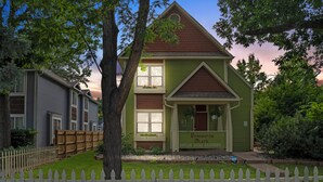 A two-story house with a green facade and a brown roof, surrounded by a white picket fence and lush greenery.
