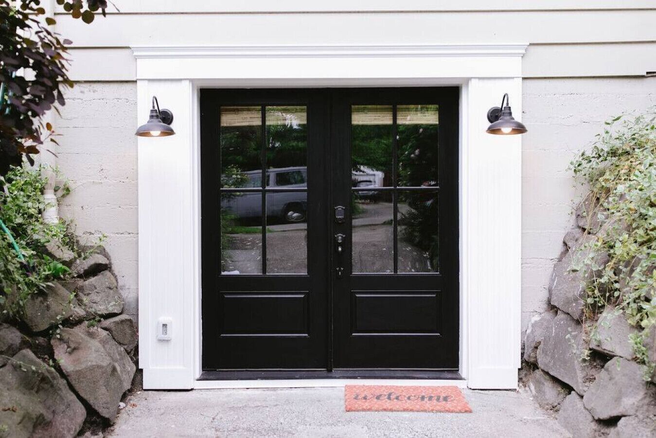 A black double door with a glass panel, a welcome mat, and two wall-mounted lights.
