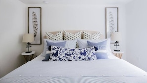 A neatly made bed with a white comforter and multiple pillows, including a distinctive blue and white patterned one. There are two bedside tables with lamps, and the room has a minimalist design with white walls and a framed artwork above the bed.