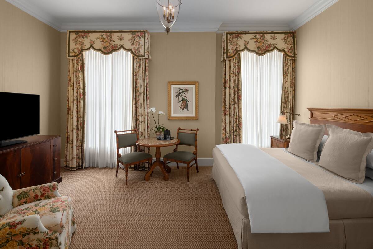 A hotel room with a bed, a desk with chairs, a television, and a painting on the wall.