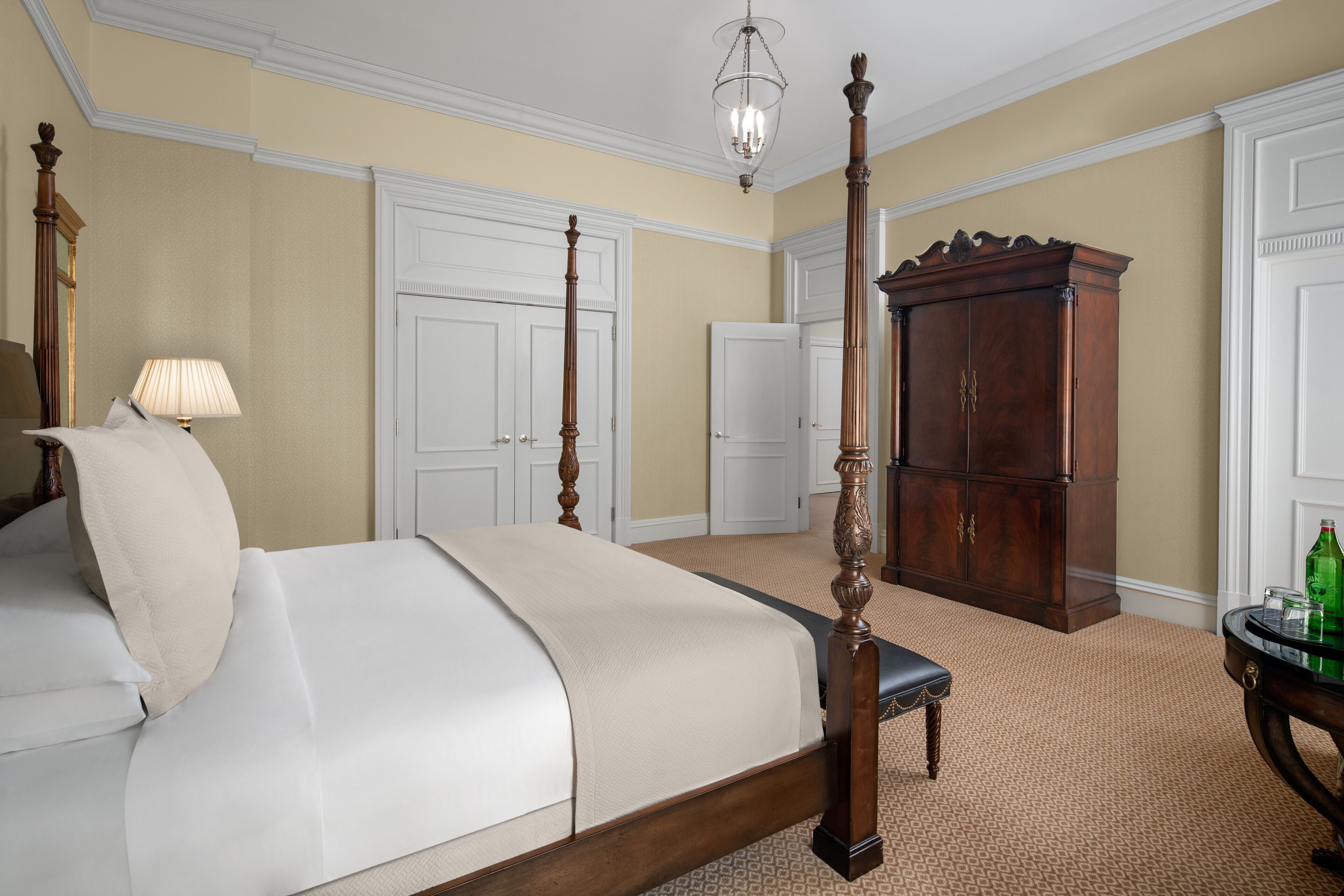 A four-poster bed with a wooden frame, a bedside table with a lamp, and a large wooden cabinet in a room with beige walls and patterned carpet.