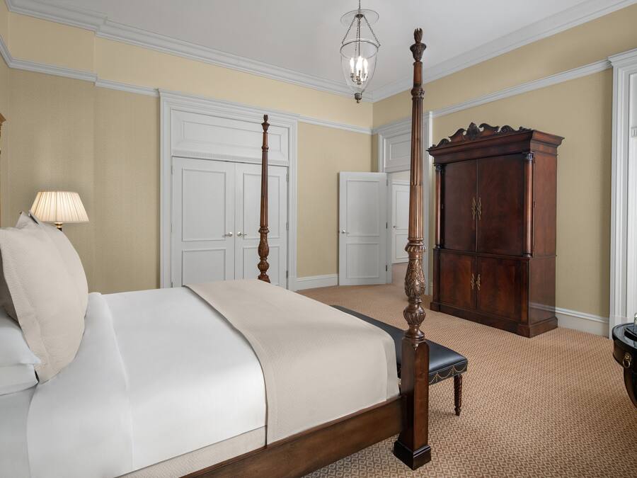 A four-poster bed with a wooden frame, a bedside table with a lamp, and a large wooden cabinet in a room with beige walls and patterned carpet.