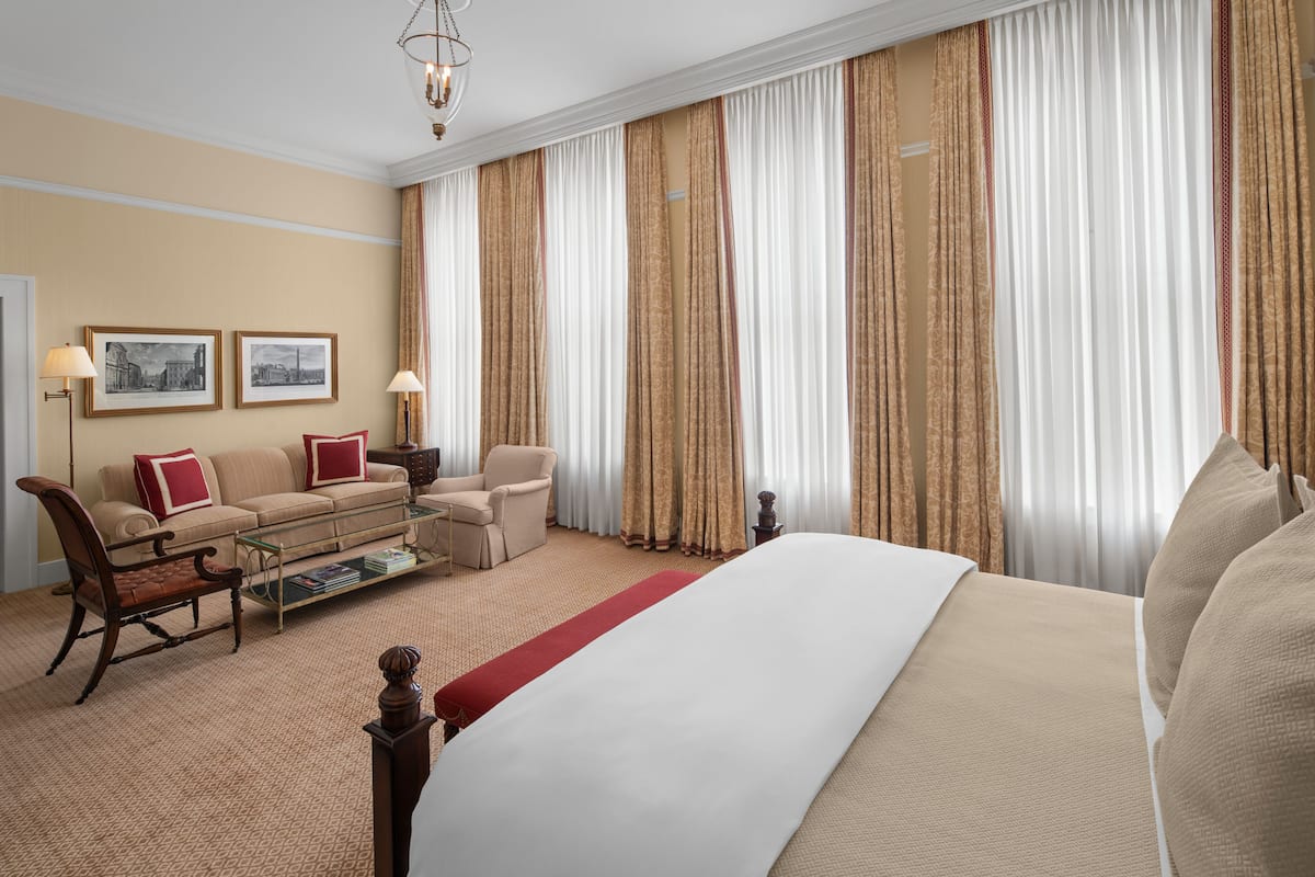 A hotel room with a large bed, a sofa, a chair, and a coffee table. There are curtains on the windows and two framed pictures on the wall.