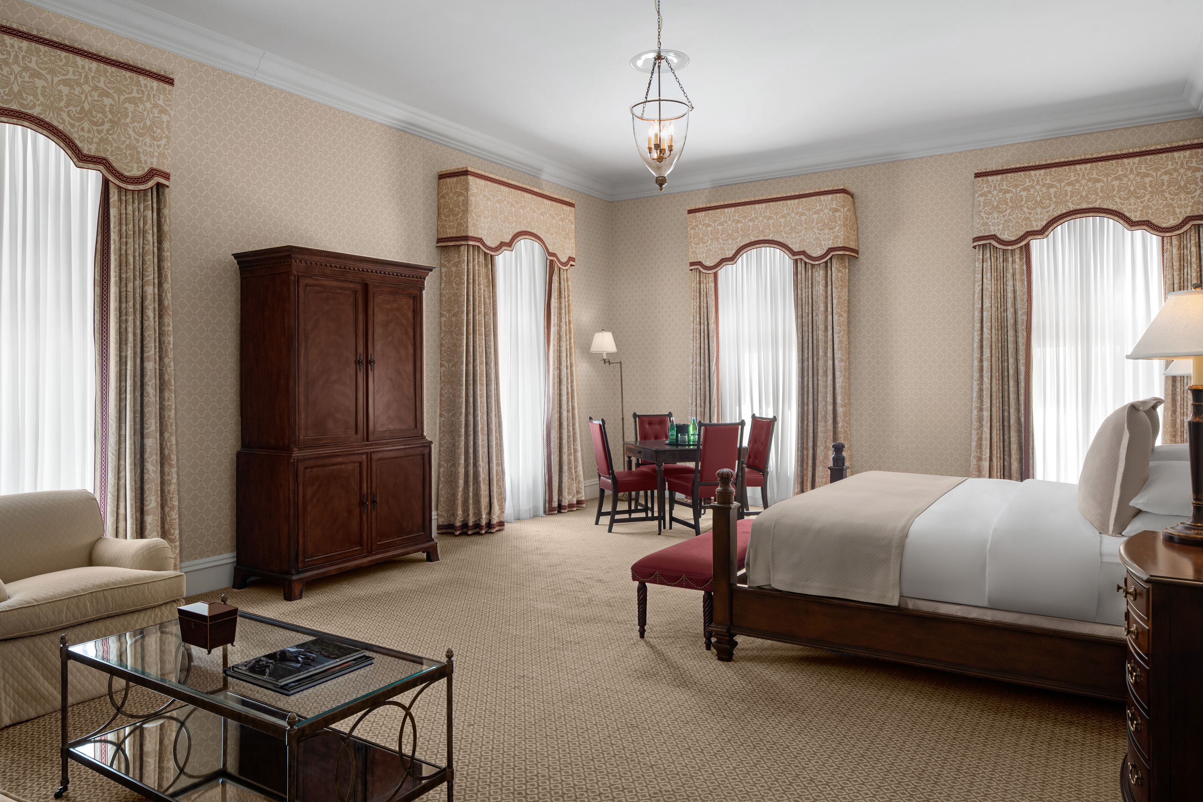Une chambre d’hôtel avec un grand lit, une armoire en bois, une table basse en verre, un bureau avec des chaises et un lustre.