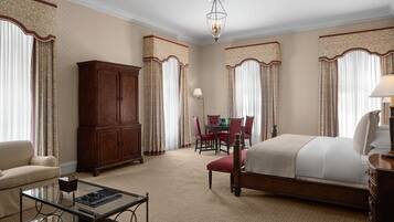 Une chambre d’hôtel avec un grand lit, une armoire en bois, une table basse en verre, un bureau avec des chaises et un lustre.