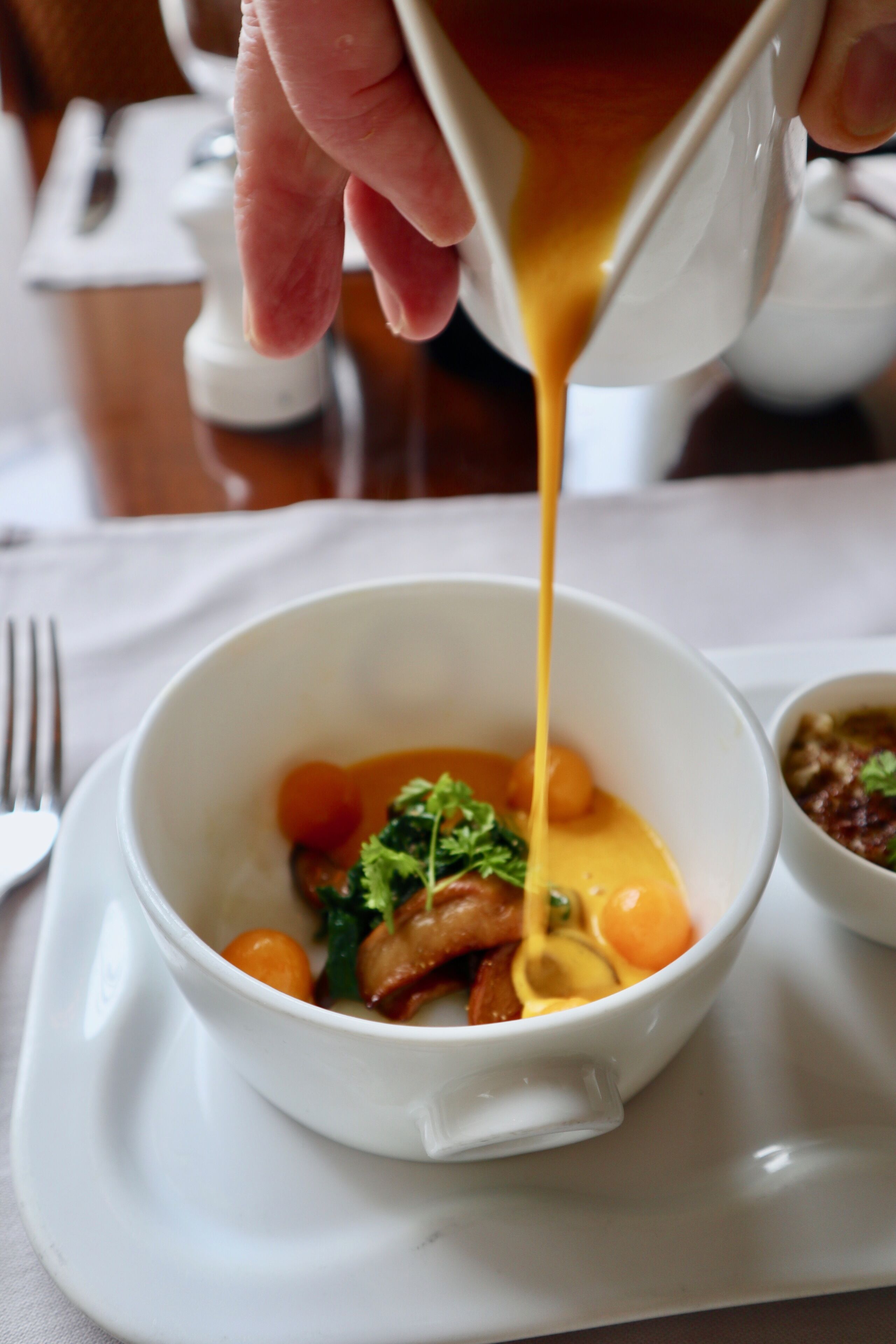 A white bowl on a white plate with a hand pouring a yellow sauce over food.
