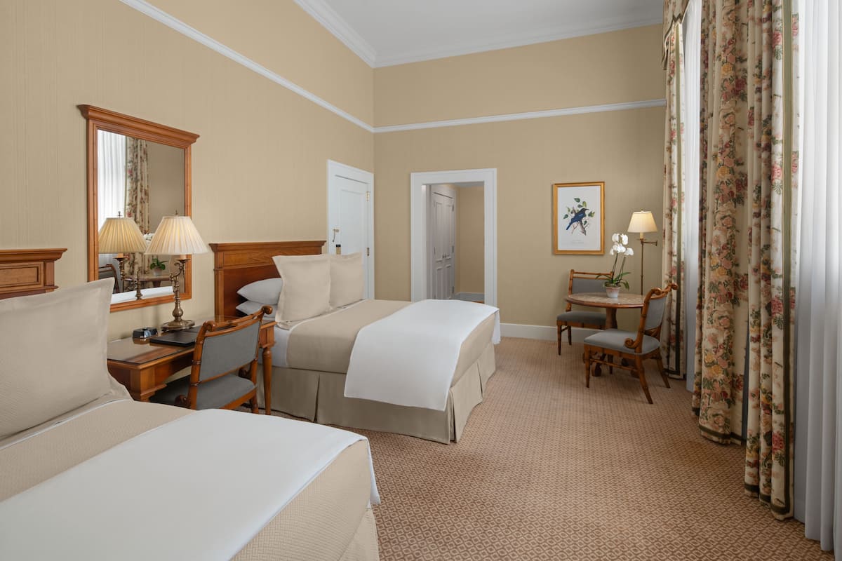 A hotel room with two beds, a desk, a chair, a small table with a lamp, a mirror, and a framed picture on the wall.
