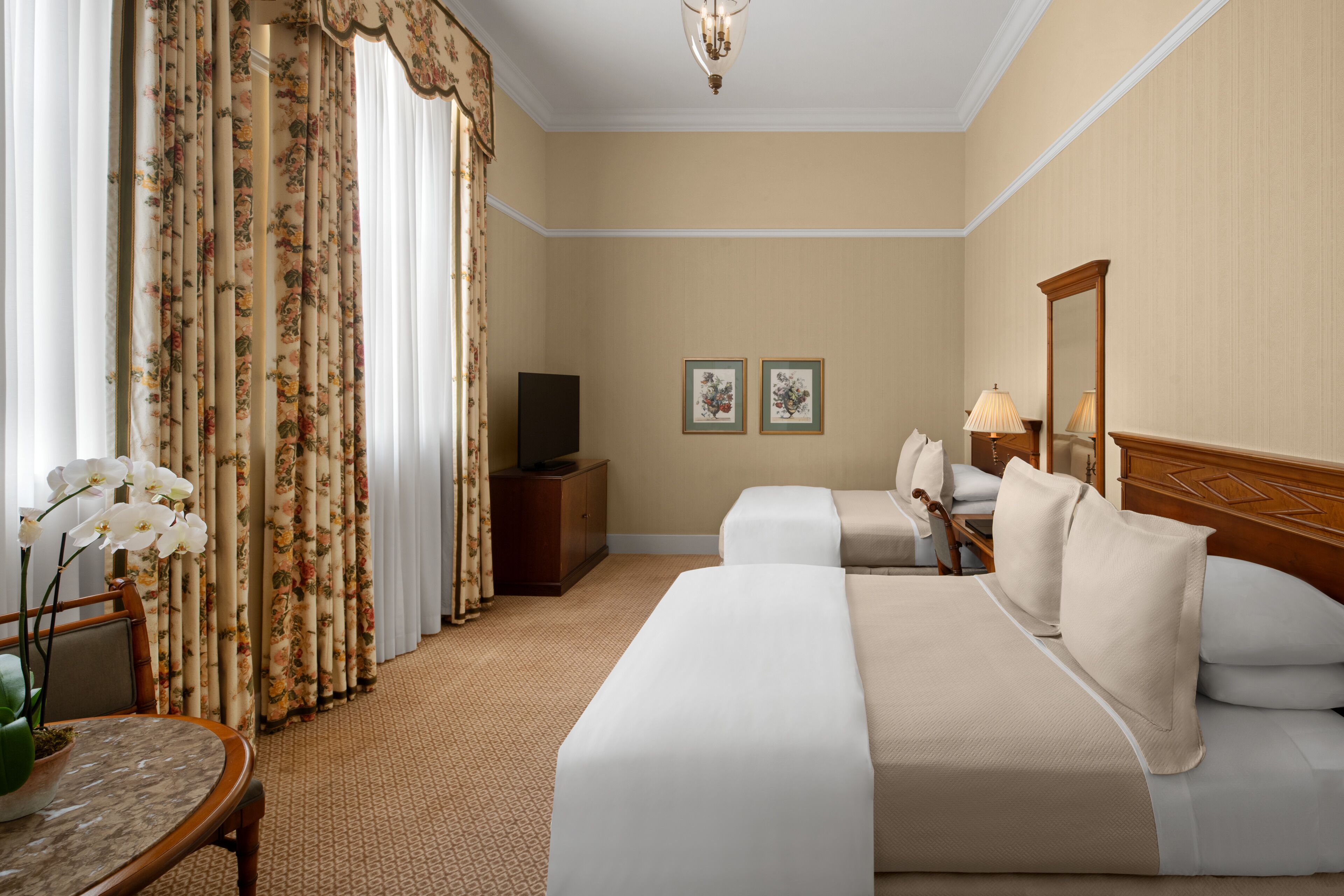 Ein Hotelzimmer mit zwei Betten, einem Fernseher auf einer Holzkommode, einem Tisch mit Blumenvase und zwei gerahmten Bildern an der Wand.