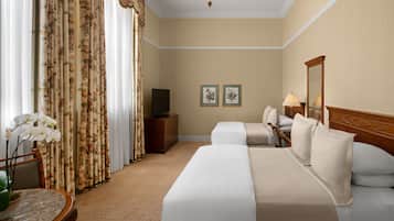 Ein Hotelzimmer mit zwei Betten, einem Fernseher auf einer Holzkommode, einem Tisch mit Blumenvase und zwei gerahmten Bildern an der Wand.