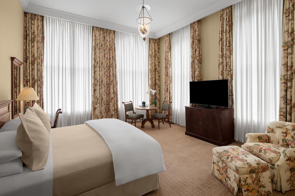 A hotel room with a bed, a TV, a desk with chairs, and floral curtains.