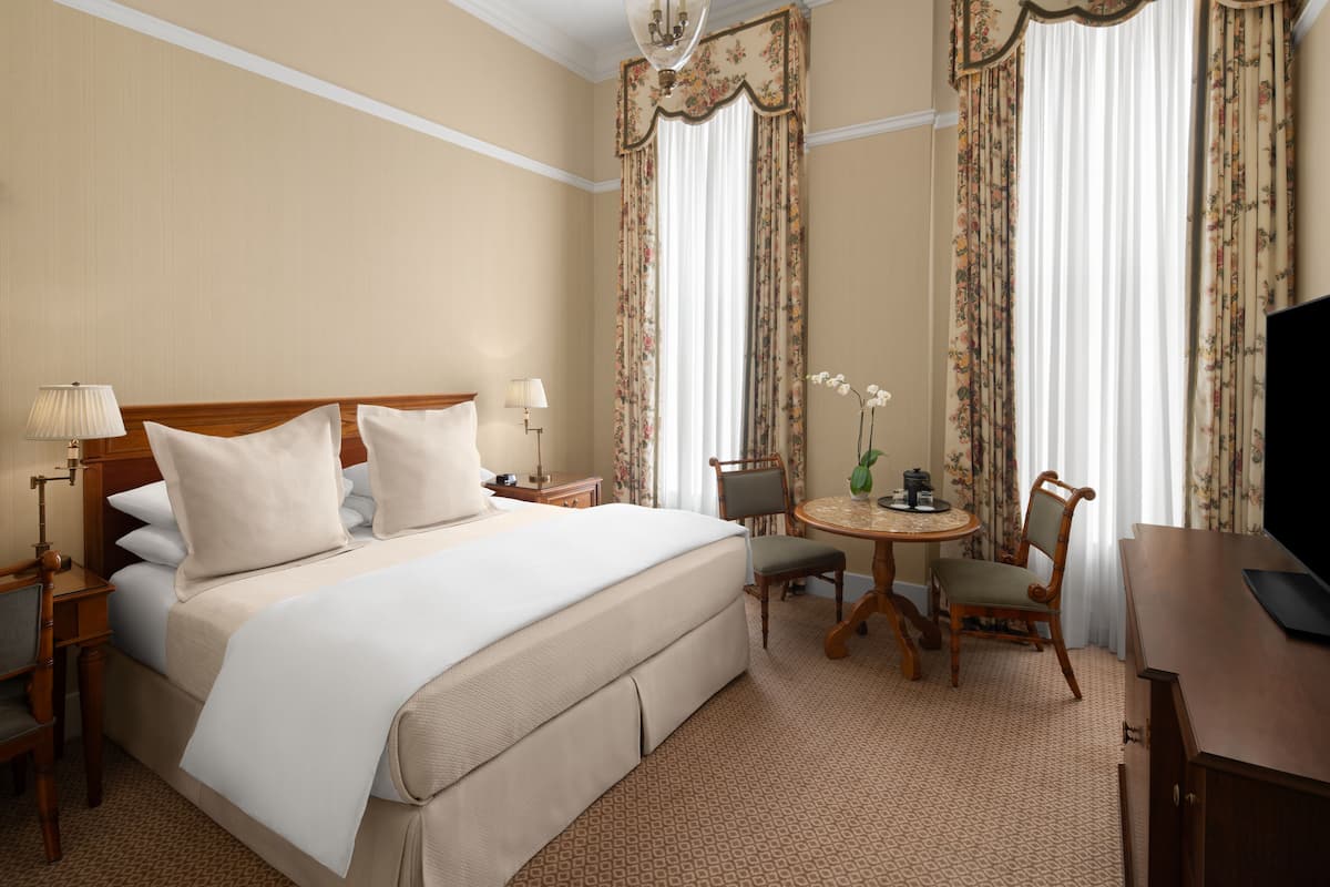 A hotel room with a large bed, a desk, a chair, a small table, and a television.