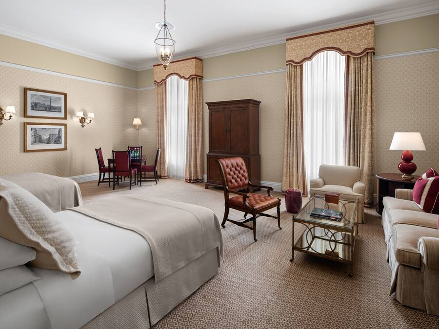 Quarto de hotel com uma cama grande, duas poltronas, um sofá, um armário de madeira e uma mesa de centro de vidro.