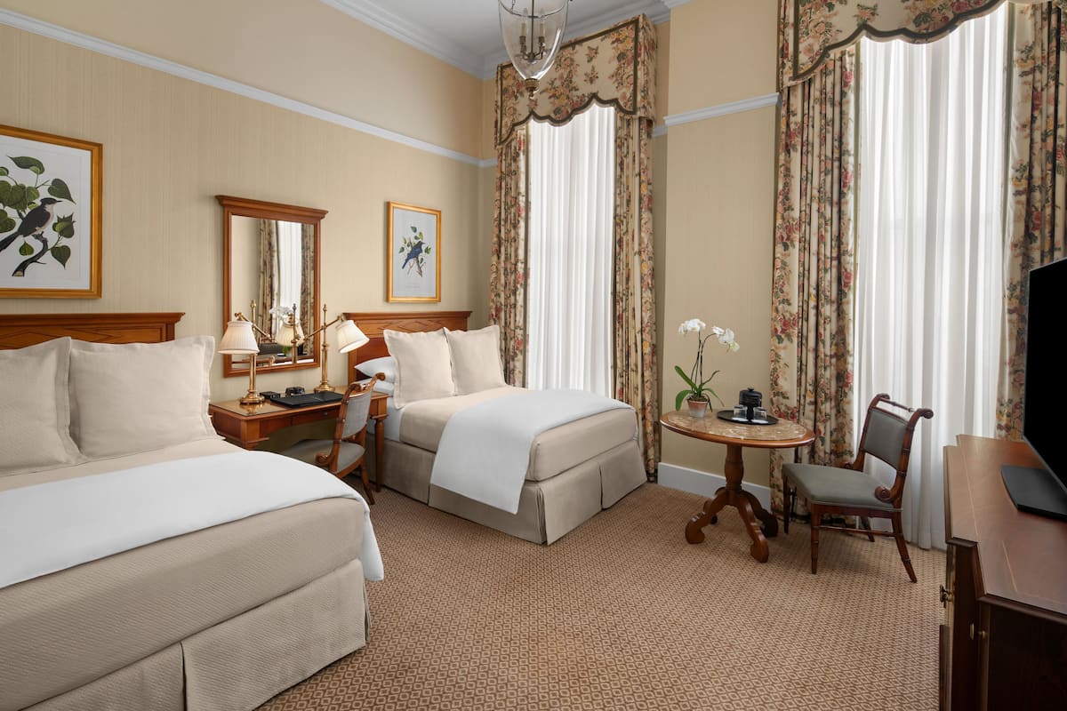 A hotel room with two beds, a desk, a chair, a television, and floral curtains.
