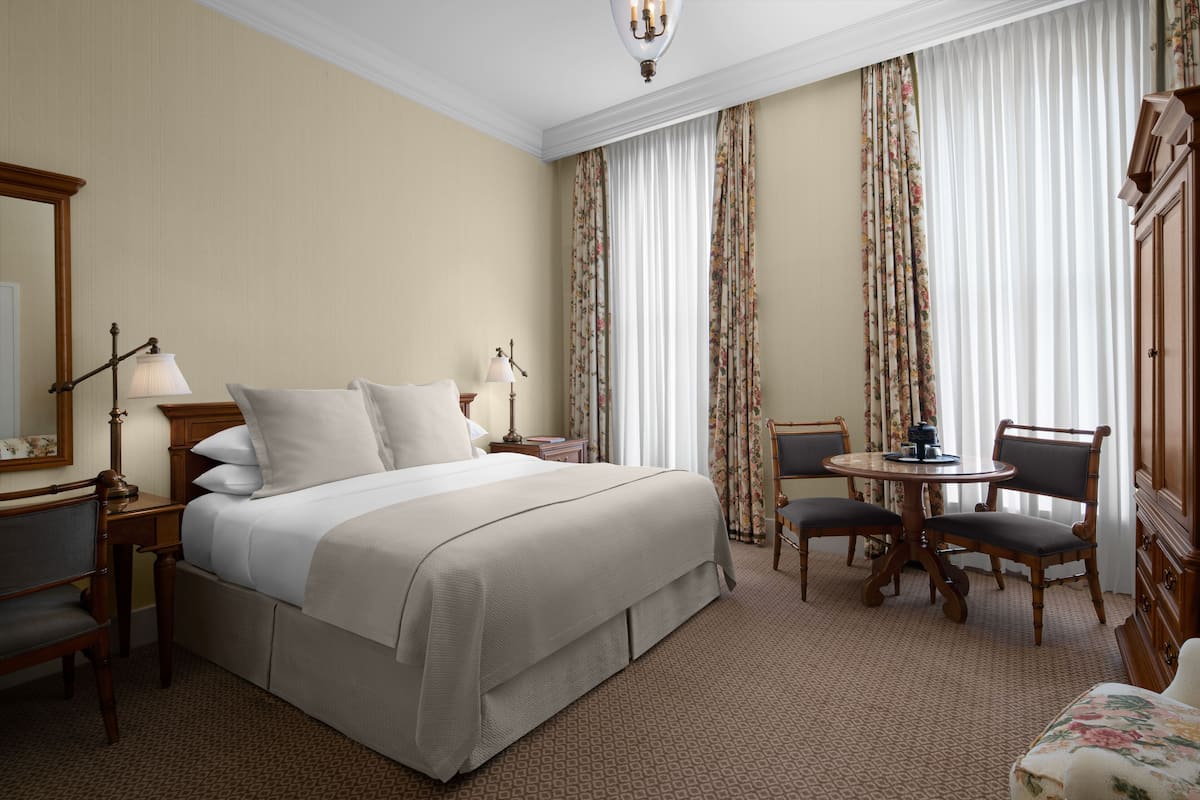 A hotel room with a large bed, a desk with a lamp, a chair, a small table, and curtains.