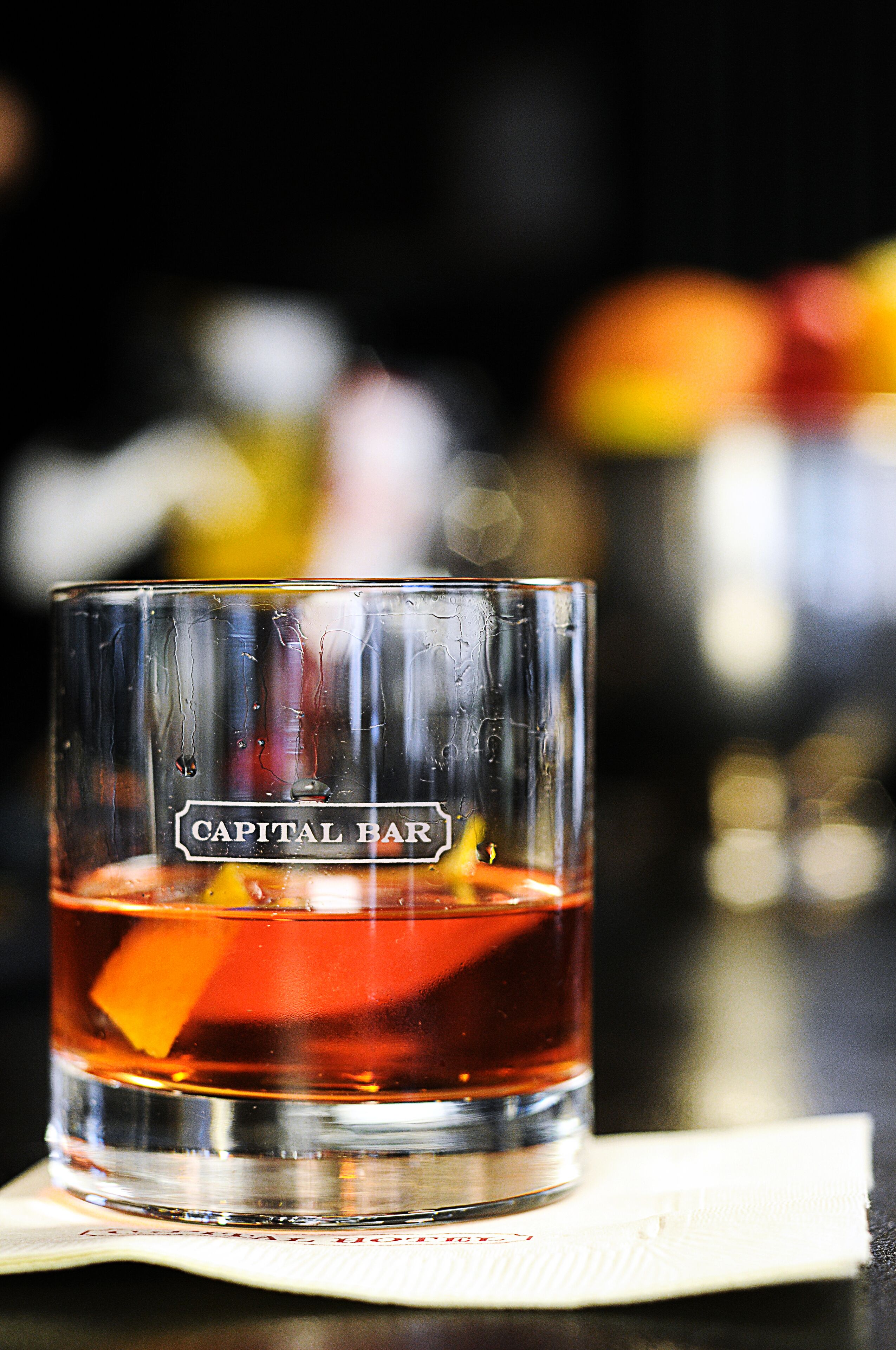 A glass of amber-colored liquid with a label reading "Capital Bar" on a bar counter.