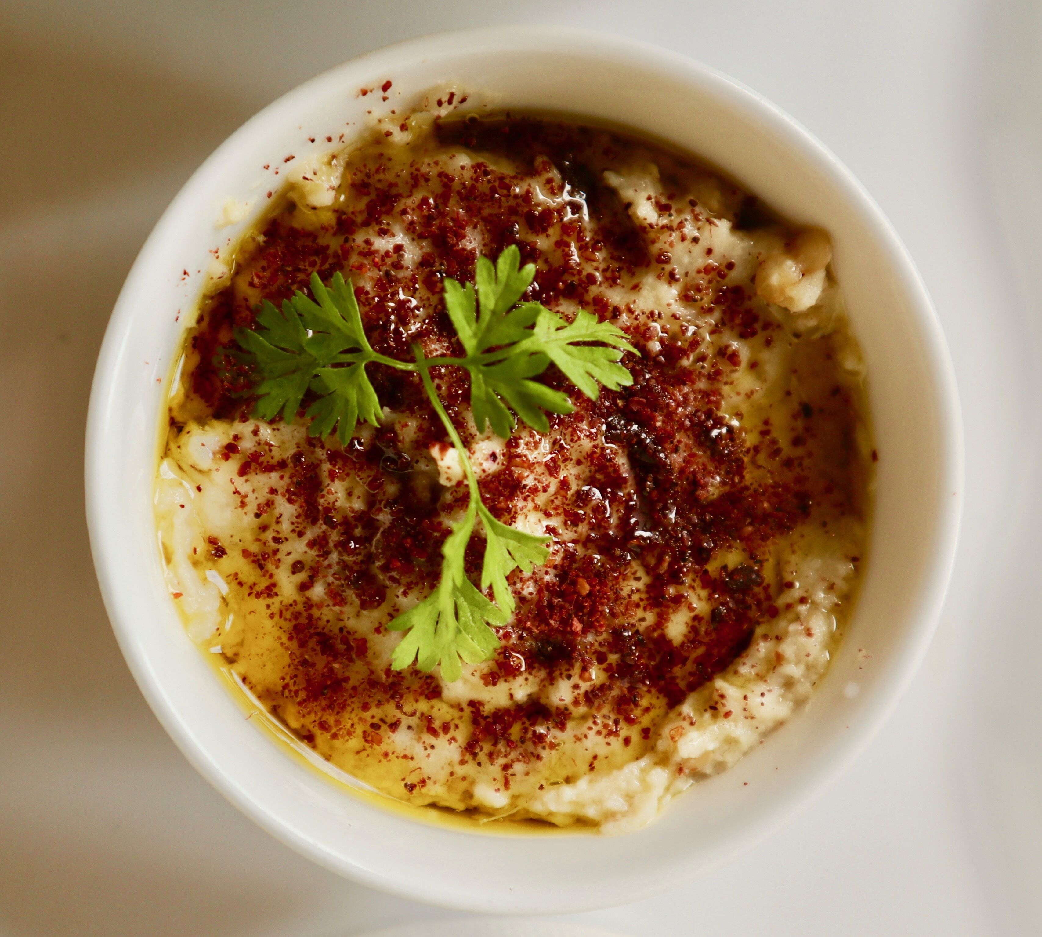 A bowl of creamy dip with a garnish of parsley and a sprinkle of red spice.