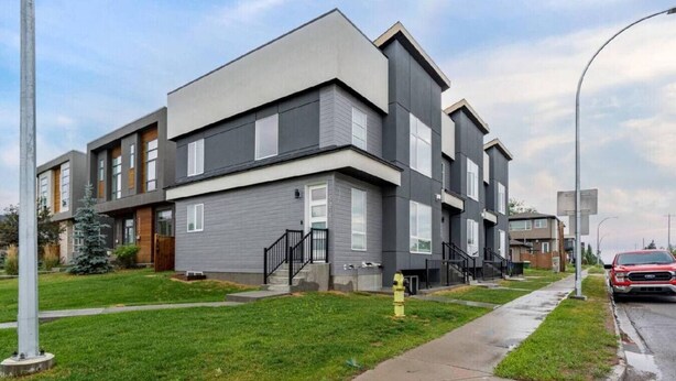 A modern multi-story residential building with a flat roof, a visible entrance with a staircase, and a well-maintained lawn.