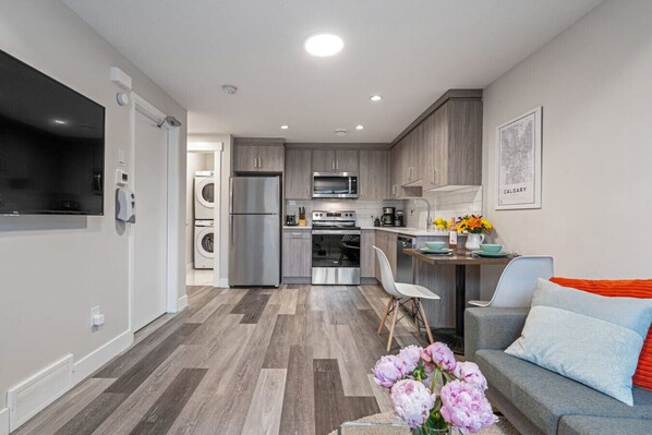 Una cocina moderna con electrodomésticos de acero inoxidable, un comedor con mesa y sillas, y una sala de estar con sofá y flores decorativas.
