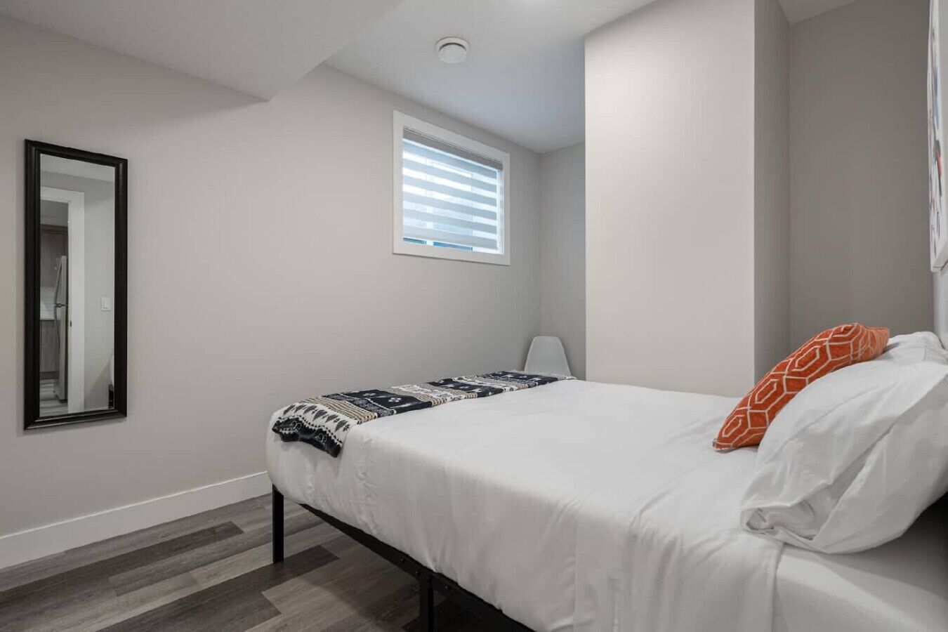 A bedroom with a bed, a window with blinds, a mirror, and a lamp.