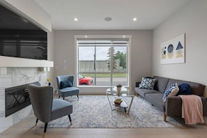 A modern living room with a fireplace, a sofa, a chair, a coffee table, and a large window with a view of a red car and trees.