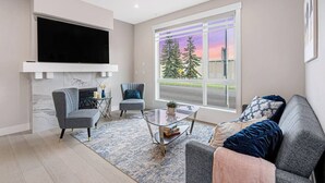 A modern living room with a fireplace, a large TV, comfortable seating, and a view of trees and a sunset through the window. - Modern 3Br Altadore Home Near Downtown (Calgary)