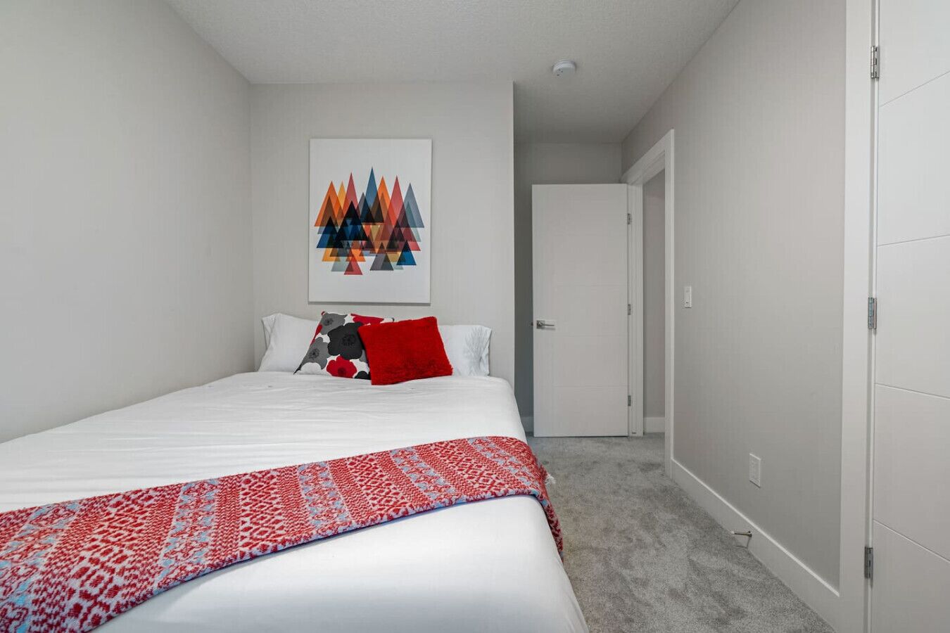 Un dormitorio con una cama, un tapete de diseño rojo y blanco, una obra de arte geométrico en la pared y una puerta cerrada.
