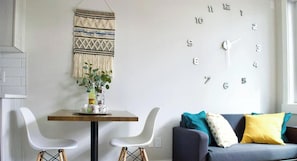 A modern living space with a wall clock, a woven tapestry, a dining table with chairs, and a sofa with cushions.