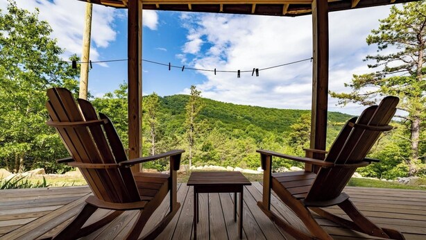 Una terraza de madera con dos mecedoras y una mesita, con vista a una ladera boscosa.