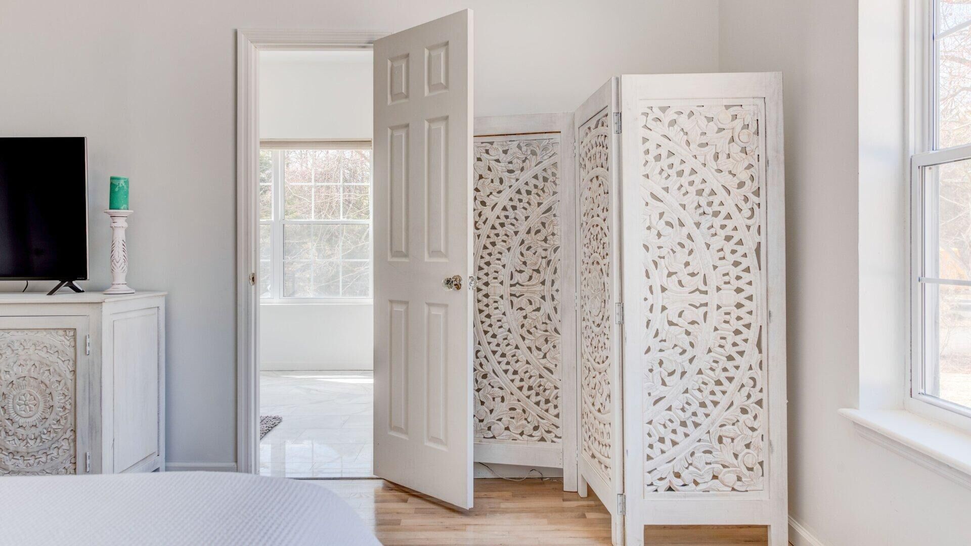 A bedroom with a bed, a TV on a cabinet, a white door, and a view through the door.