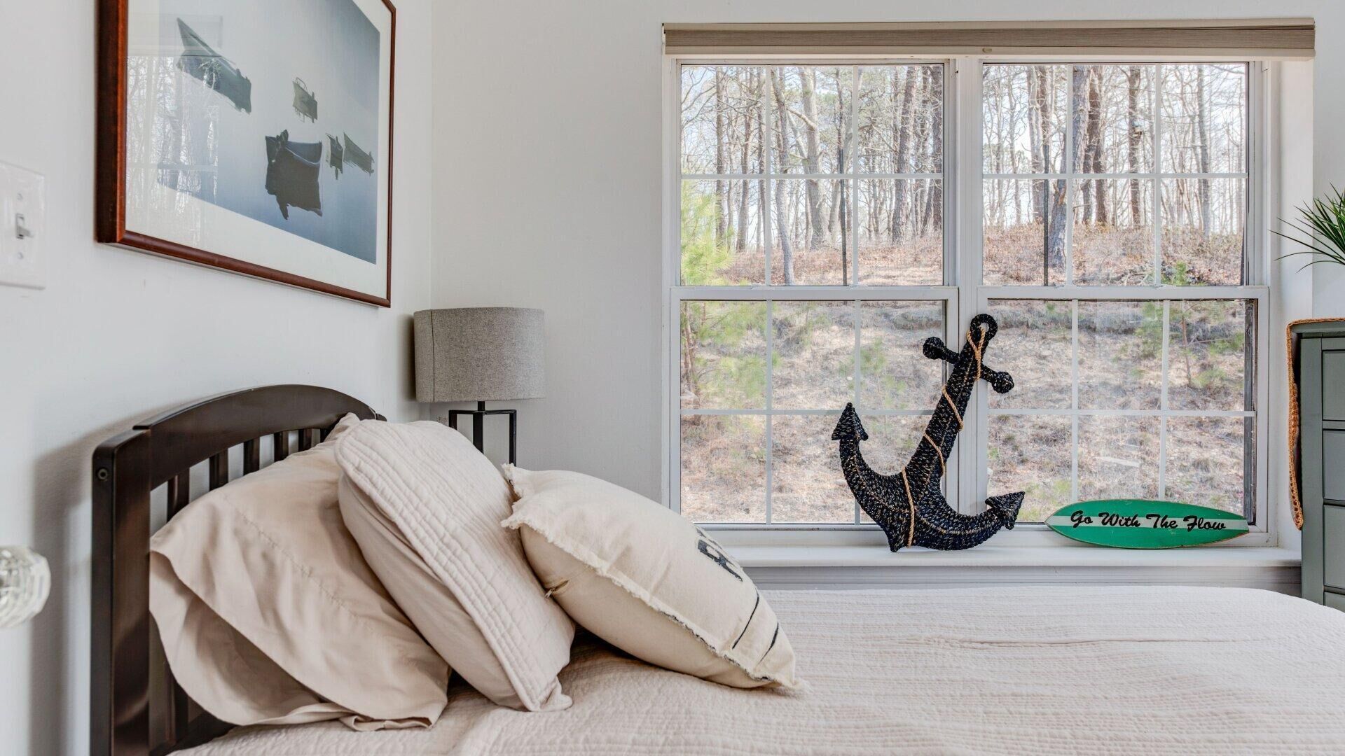 A bedroom with a large window, a bed with pillows, a lamp, a framed picture, and a decorative anchor.