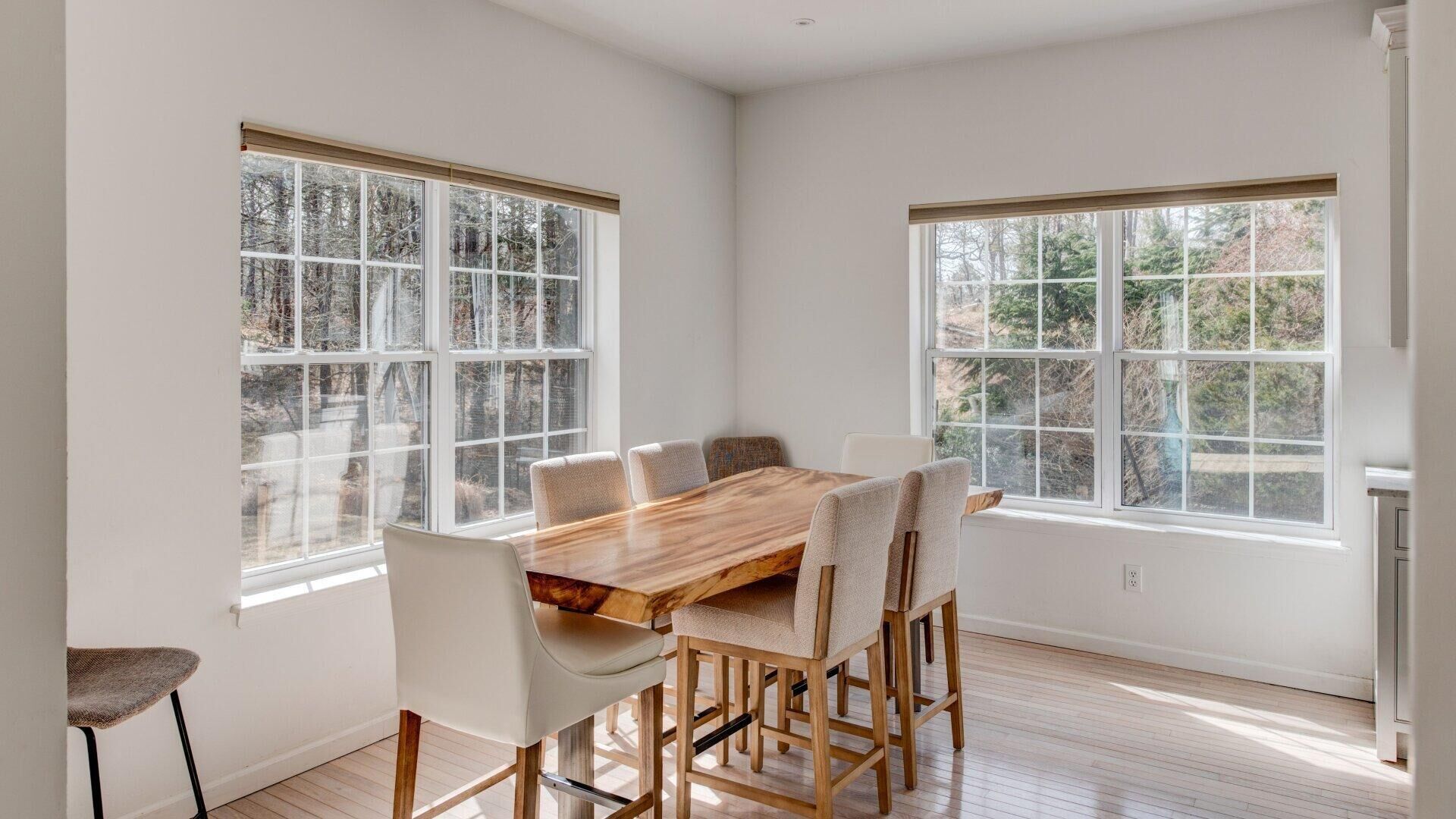 A dining area with a wooden table and chairs, large windows with a view of trees, and a white interior.