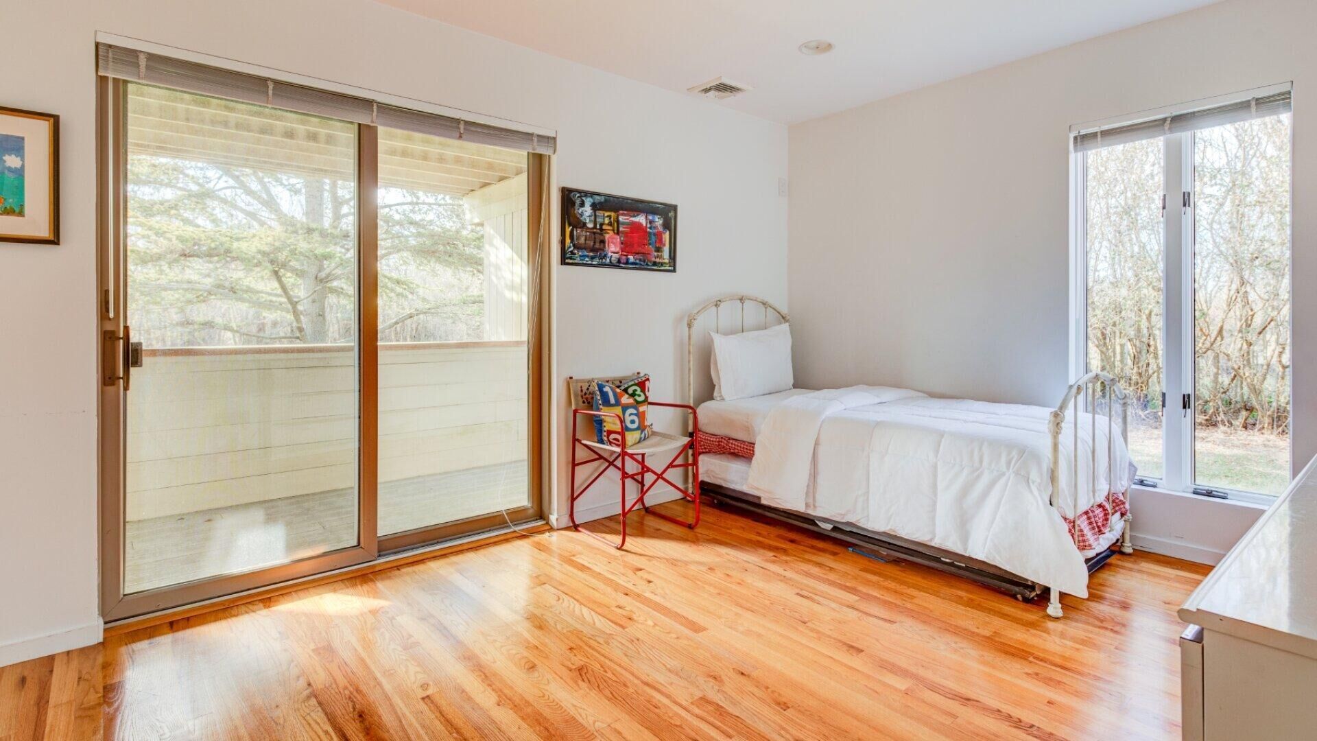 Un dormitorio con piso de madera, una cama con ropa blanca, una silla roja y un ventanal grande.