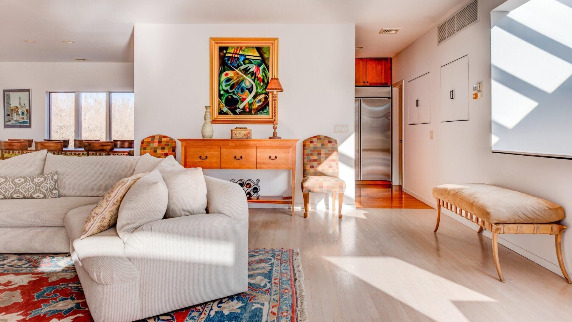 A spacious living room with a sofa, armchairs, a coffee table, and a rug. There is a painting on the wall, a dining area in the background, and a kitchen visible through an open door.