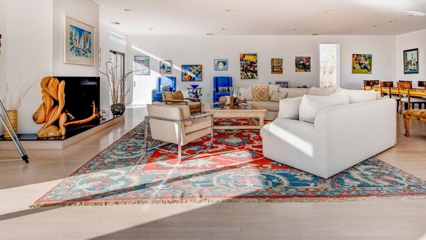 A modern living room with a white sofa, a coffee table, and a large patterned rug. There are abstract paintings on the walls and a fireplace with a sculpture. - Bridgehampton Beach Walk (Bridgehampton)