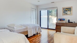 A bedroom with two beds, a sliding glass door to a balcony, a sofa, and a small cabinet with artwork.
