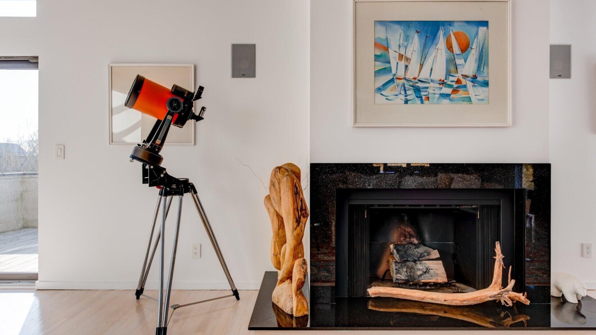 A room with a telescope on a tripod, a fireplace with logs, and a framed painting of sailboats.