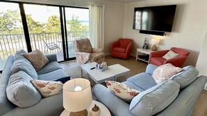 A modern living room with a grey sofa set, a small white table with a lamp, and a red armchair. A flat-screen TV is mounted on the wall. - Newly renovated Oceanside condo with easy access to the pool and beach! (Ocean City)