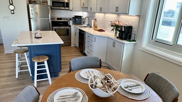 A modern kitchen with white cabinets, a central island, and a dining area with a wooden table set for four. - Newly renovated Oceanside condo with easy access to the pool and beach! (Ocean City)