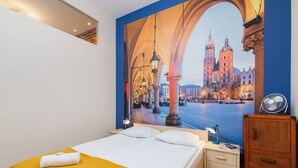 Une chambre d’hôtel avec une grande fenêtre, un lit avec du linge de maison blanc, une table de chevet en bois et un ventilateur.