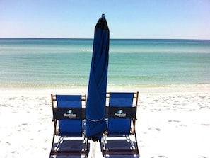 Zwei blaue Liegestühle mit einem blauen Sonnenschirm an einem sandigen Strand, mit dem Meer im Hintergrund.