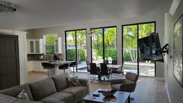 Ein modernes Wohnzimmer mit einer Couch, einem Couchtisch und einem Fernseher. Es hat große Fenster mit Blick auf Grünflächen und eine Terrasse mit Sitzgelegenheiten.