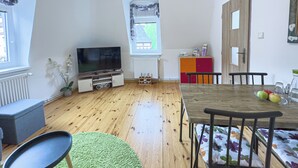Ein Wohnzimmer mit Holzboden, einem Fernsehgerät auf einem Schrank, einem Esstisch mit Stühlen und einem Fenster mit Vorhängen.