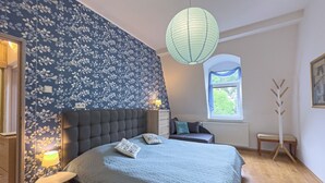 Ein Schlafzimmer mit einem gepolsterten Kopfteil, einem Bett mit zwei Kissen, einem Nachttisch mit Lampe, einem Fenster mit Blick und einer stehenden Stehlampe.
