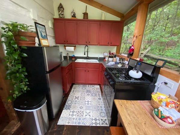 A compact kitchen with red cabinets, a stainless steel refrigerator, a gas stove, and a window with a view of greenery.