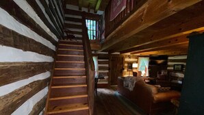 A log cabin interior with a wooden staircase, a living area with a sofa and a fireplace, and a window with curtains.